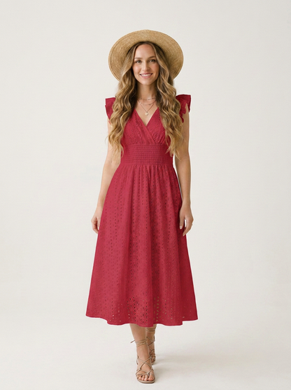 A woman modeling a sleeveless red eyelet midi dress with a V-neckline and flared skirt, styled with a wide-brimmed straw hat and strappy sandals against a plain light background.