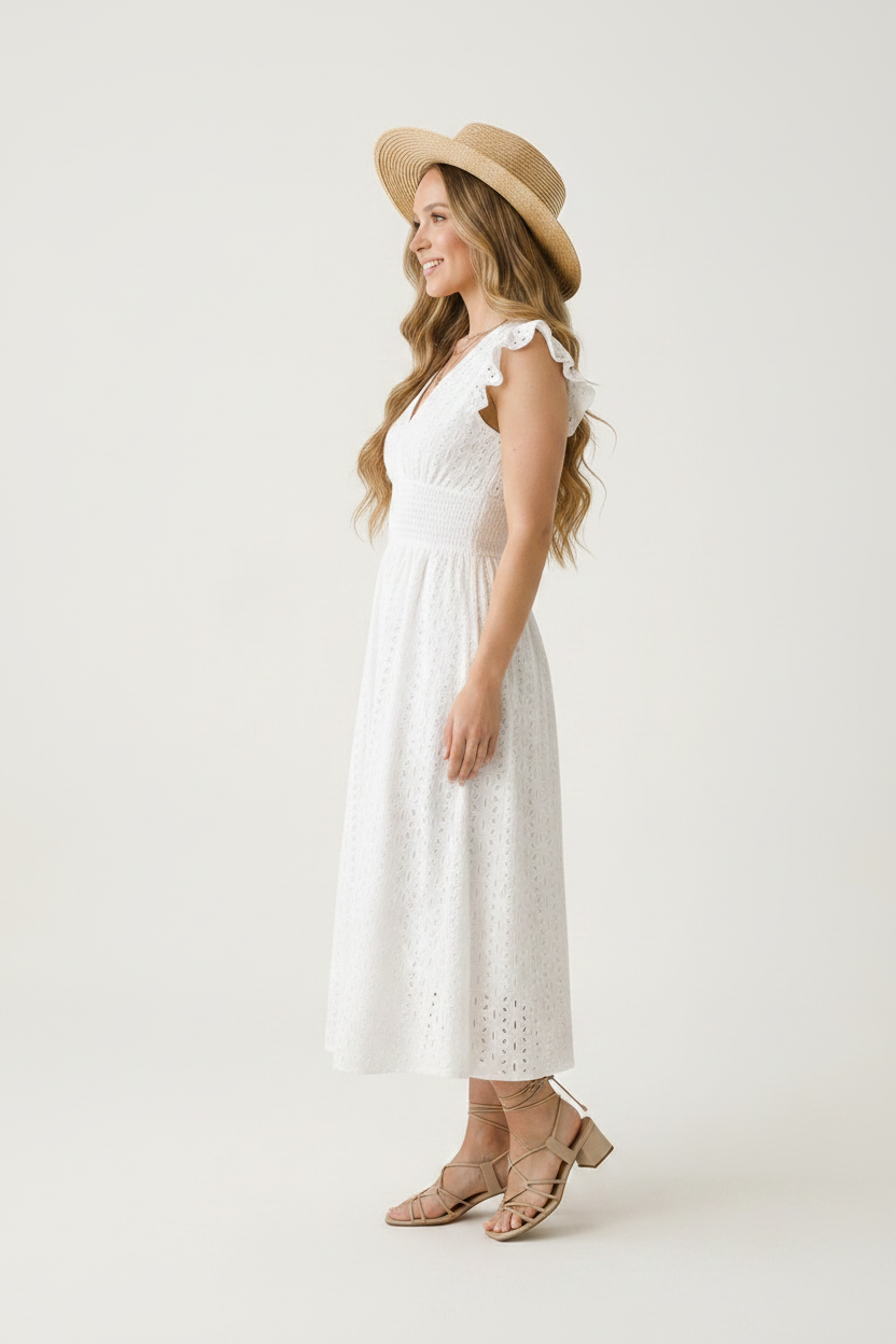 A woman modeling a white eyelet midi dress with ruffled cap sleeves and a V-neckline, paired with a wide-brimmed straw hat and tan strappy heels, against a plain light background.