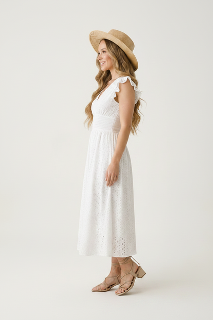 A woman modeling a white eyelet midi dress with ruffled cap sleeves and a V-neckline, paired with a wide-brimmed straw hat and tan strappy heels, against a plain light background.