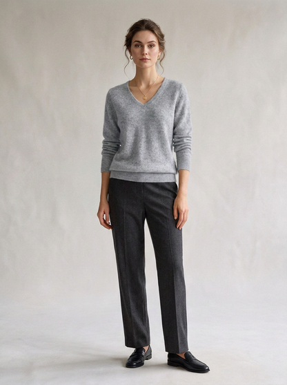 A woman modeling a light gray V-neck knit sweater and dark charcoal gray trousers, paired with black loafers, against a neutral light gray studio backdrop.