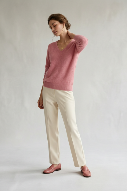 A woman modeling a dusty pink knit V-neck sweater, paired with cream-colored trousers and matching pink leather loafers, against a neutral light gray studio background.