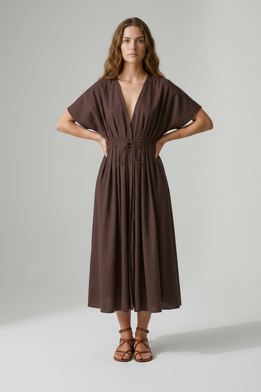 A woman modeling a dark brown midi wrap dress with short flutter sleeves, a deep V-neckline, and a drawstring waist, paired with brown strappy sandals against a neutral studio background.