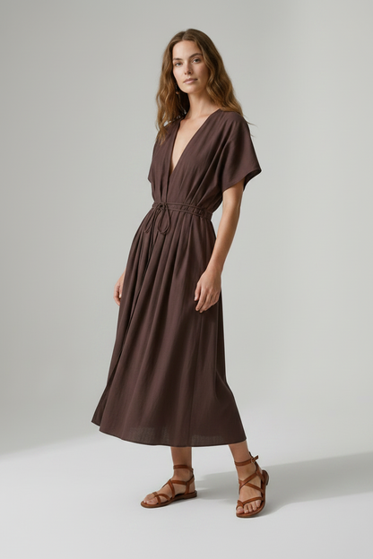 A woman modeling a dark brown midi wrap dress with short flutter sleeves, a deep V-neckline, and a drawstring waist, paired with brown strappy sandals against a neutral studio background.