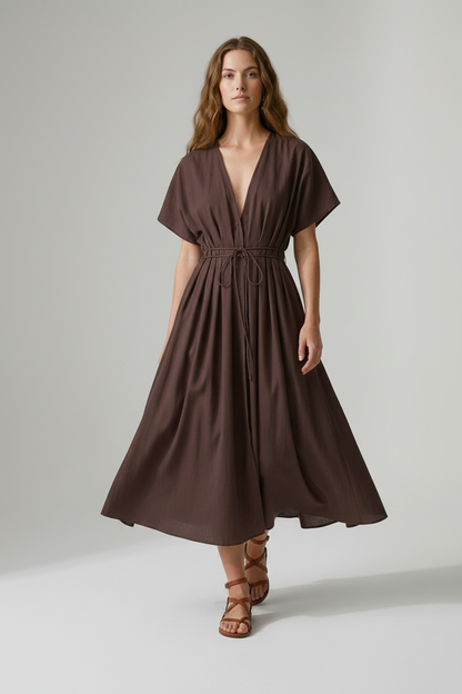 A woman modeling a dark brown midi wrap dress with short flutter sleeves, a deep V-neckline, and a drawstring waist, paired with brown strappy sandals against a neutral studio background.