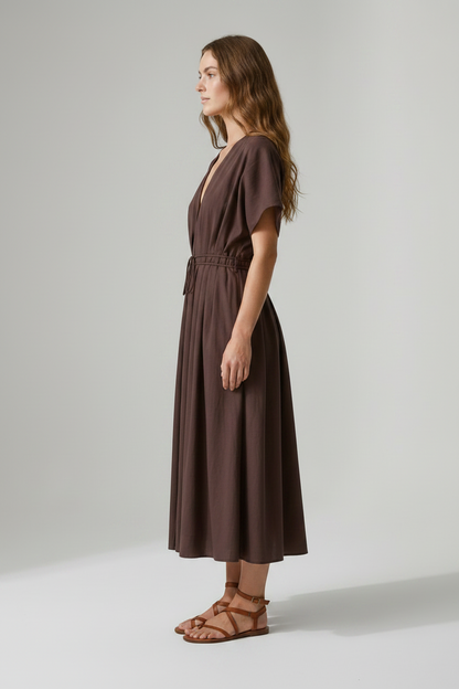 A woman modeling a dark brown midi wrap dress with short flutter sleeves, a deep V-neckline, and a drawstring waist, paired with brown strappy sandals against a neutral studio background.