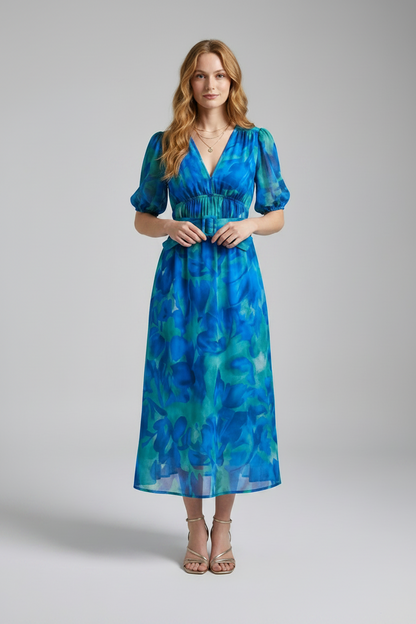 A woman modeling a midi-length blue and green abstract print dress with puff sleeves, a V-neckline, and a smocked waist, set against a plain gray studio background.