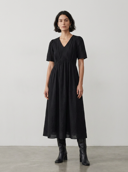 A woman modeling a black midi dress with short puff sleeves, a V-neckline, and a gathered waist, paired with black ankle boots against a neutral studio backdrop.