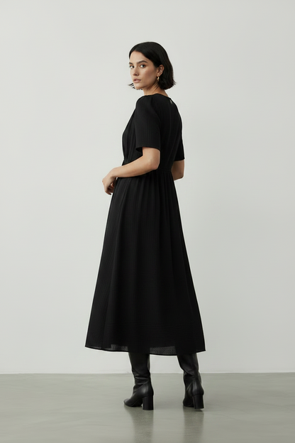 A woman modeling a black midi dress with short sleeves, a V-neckline, and a gathered waist, paired with black ankle boots, set against a minimalist light gray studio background.