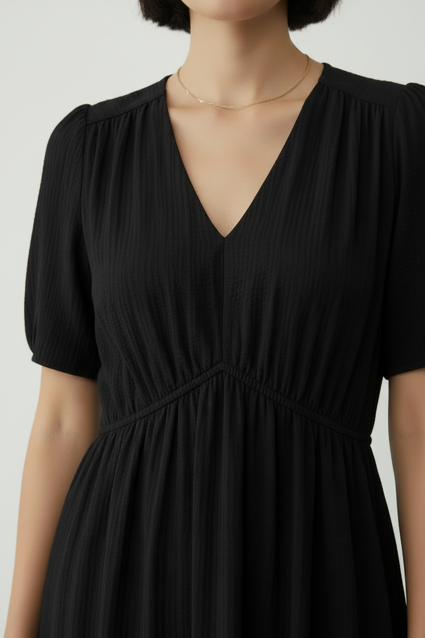 A close-up of a model wearing a black short-sleeve V-neck dress with a textured crinkle fabric, paired with a delicate gold chain necklace.