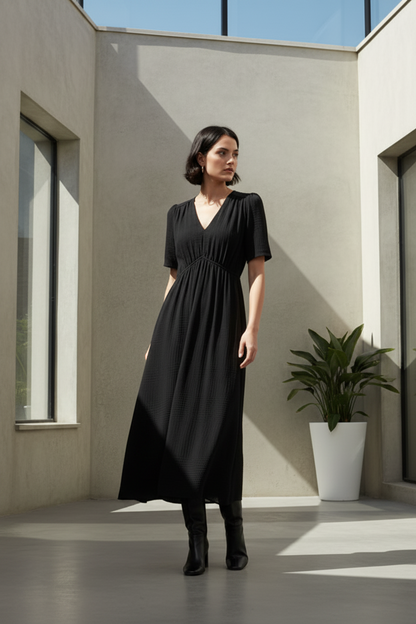 A woman stands in a modern concrete courtyard wearing a black midi dress with short sleeves, a V-neckline, and a gathered waist, paired with black boots.