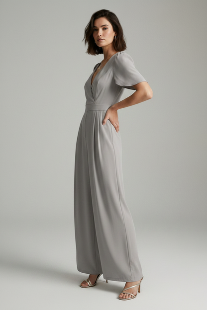 A woman modeling a light gray jumpsuit with short puff sleeves, a wrap-style V-neck bodice, and wide legs, photographed against a plain light gray studio background.