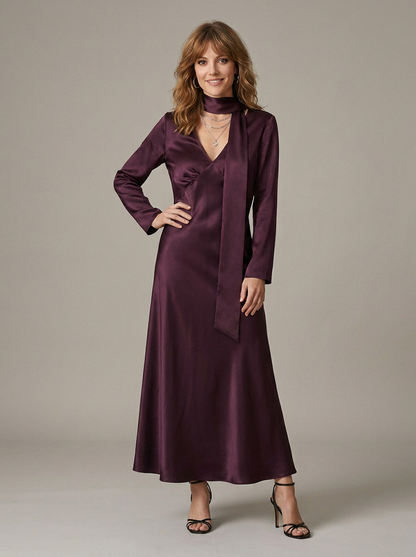 A woman models a long-sleeved, floor-length satin dress in a deep plum purple color, featuring a high neck, a wrap-style front overlay, and a V-neckline, paired with black strappy heels against a neutral studio backdrop.