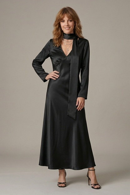 A woman models a long-sleeved, floor-length satin dress in a deep plum purple color, featuring a high neck, a wrap-style front overlay, and a V-neckline, paired with black strappy heels against a neutral studio backdrop.