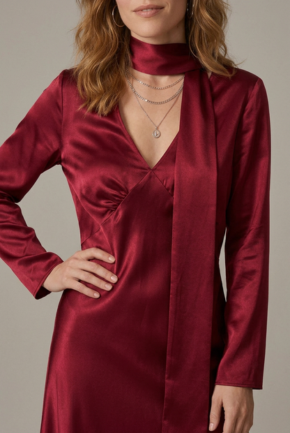A woman modeling a long-sleeved satin maxi dress in deep burgundy red, featuring a high neck, V-neckline, and a matching self-tie belt, set against a plain light gray studio background.