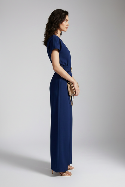 A woman models a navy blue short-sleeve jumpsuit with a gold-tone belt and decorative buckle, styled with a beige clutch and neutral sandals against a light gray studio background.