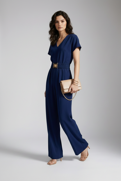 A woman models a navy blue short-sleeve jumpsuit with a gold-tone belt buckle, holding a beige clutch bag and wearing beige heels against a neutral gray studio background.