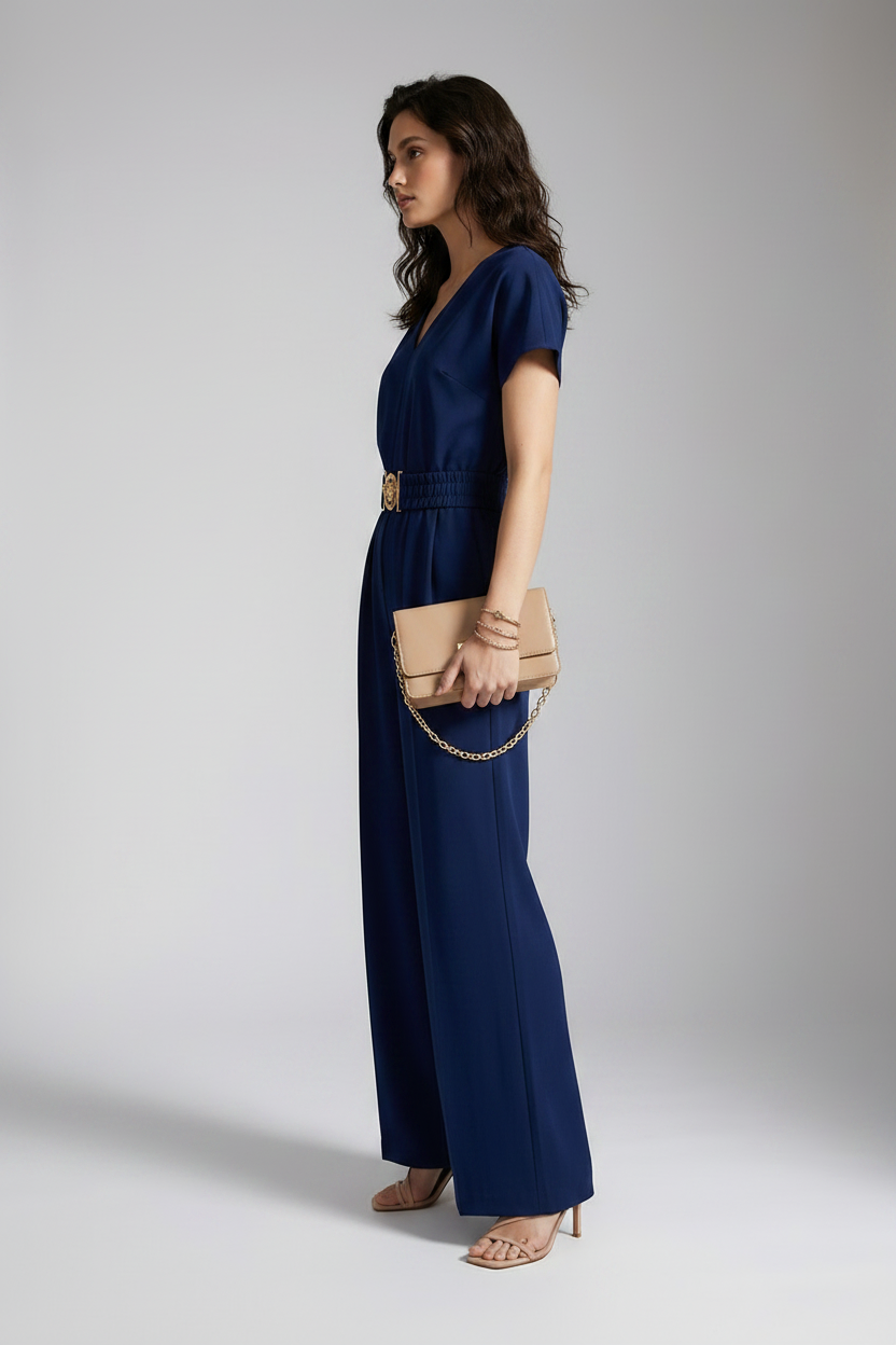 A woman models a navy blue short-sleeve jumpsuit with a gold-tone belt and decorative buckle, styled with a beige clutch and neutral sandals against a light gray studio background.