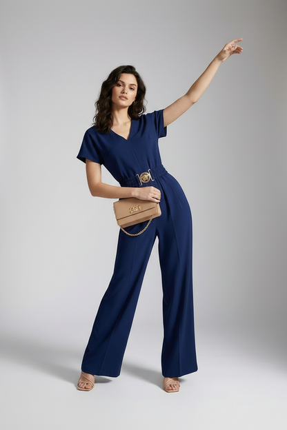 A woman models a navy blue short-sleeve jumpsuit with a gold-tone belt and decorative buckle, styled with a beige clutch and neutral sandals against a light gray studio background.