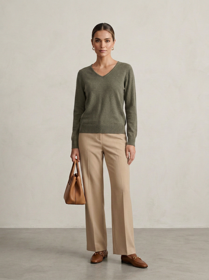 A woman stands in a studio wearing an olive green V-neck sweater, beige wide-leg trousers, brown loafers, and holding a tan leather handbag against a light textured wall.