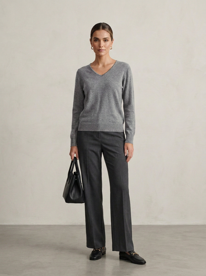 A woman stands in a studio wearing a heather gray long-sleeve V-neck sweater, dark charcoal wide-leg trousers, black loafers, and holding a black leather handbag.