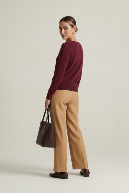 A woman stands in a studio wearing a burgundy V-neck sweater, tan wide-leg trousers, and dark brown loafers, while holding a dark brown tote bag. The background is a clean, textured light beige wall and gray floor.
