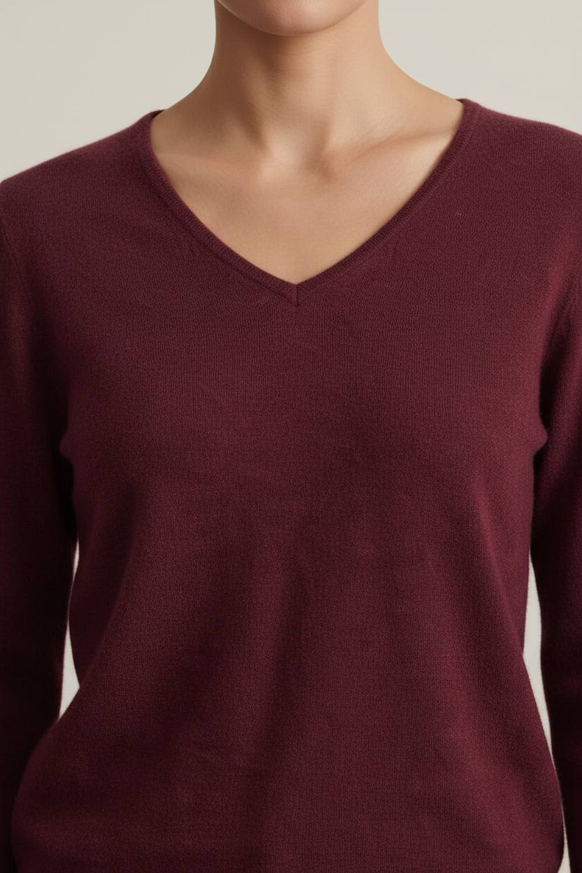 A woman stands in a studio wearing a burgundy V-neck sweater, tan wide-leg trousers, and dark brown loafers, while holding a dark brown tote bag. The background is a clean, textured light beige wall and gray floor.