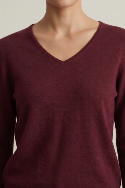 A woman stands in a studio wearing a burgundy V-neck sweater, tan wide-leg trousers, and dark brown loafers, while holding a dark brown tote bag. The background is a clean, textured light beige wall and gray floor.