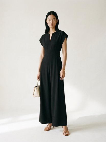 A woman stands in a studio wearing a black sleeveless maxi dress with a split neckline and wide legs, holding a small beige handbag. She is wearing tan heeled sandals against a plain light background.