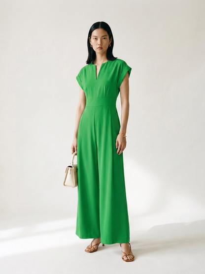A woman modeling a bright green wide-leg jumpsuit with cap sleeves and a split neckline, holding a small beige handbag against a plain light background.