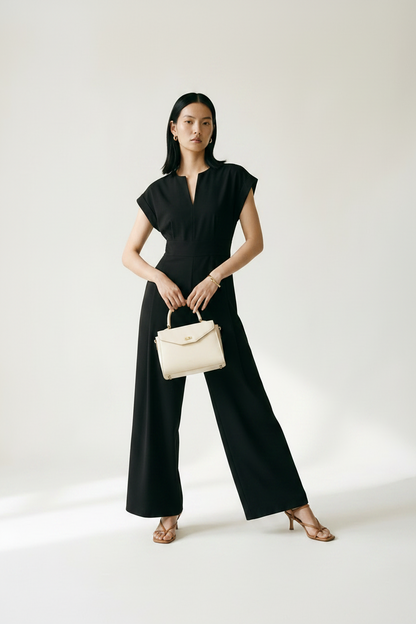 A woman stands in a studio wearing a black sleeveless maxi dress with a split neckline and wide legs, holding a small beige handbag. She is wearing tan heeled sandals against a plain light background.