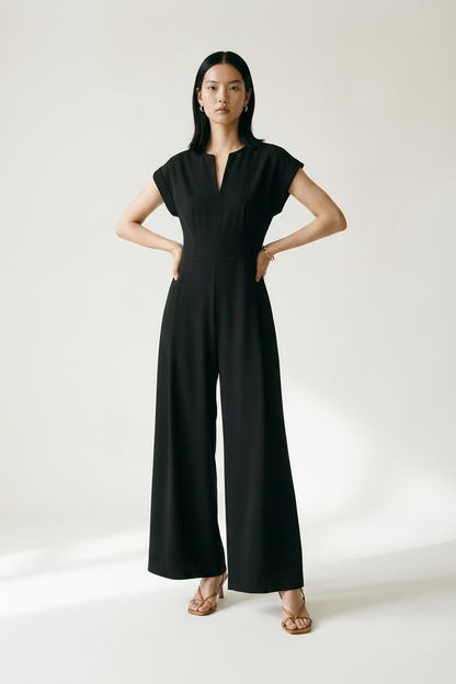A woman stands in a studio wearing a black sleeveless maxi dress with a split neckline and wide legs, holding a small beige handbag. She is wearing tan heeled sandals against a plain light background.