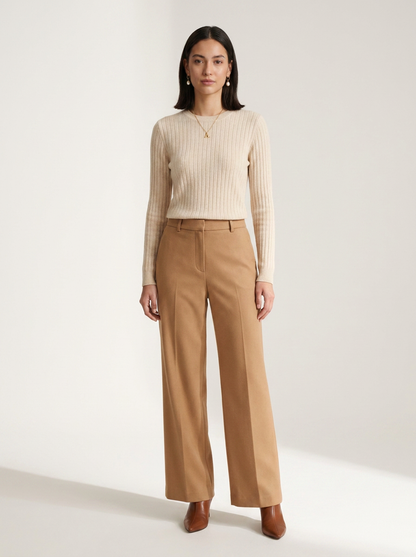 A woman stands against a plain light beige studio background wearing a cream ribbed long-sleeve top and tan wide-leg trousers with brown pointed shoes.