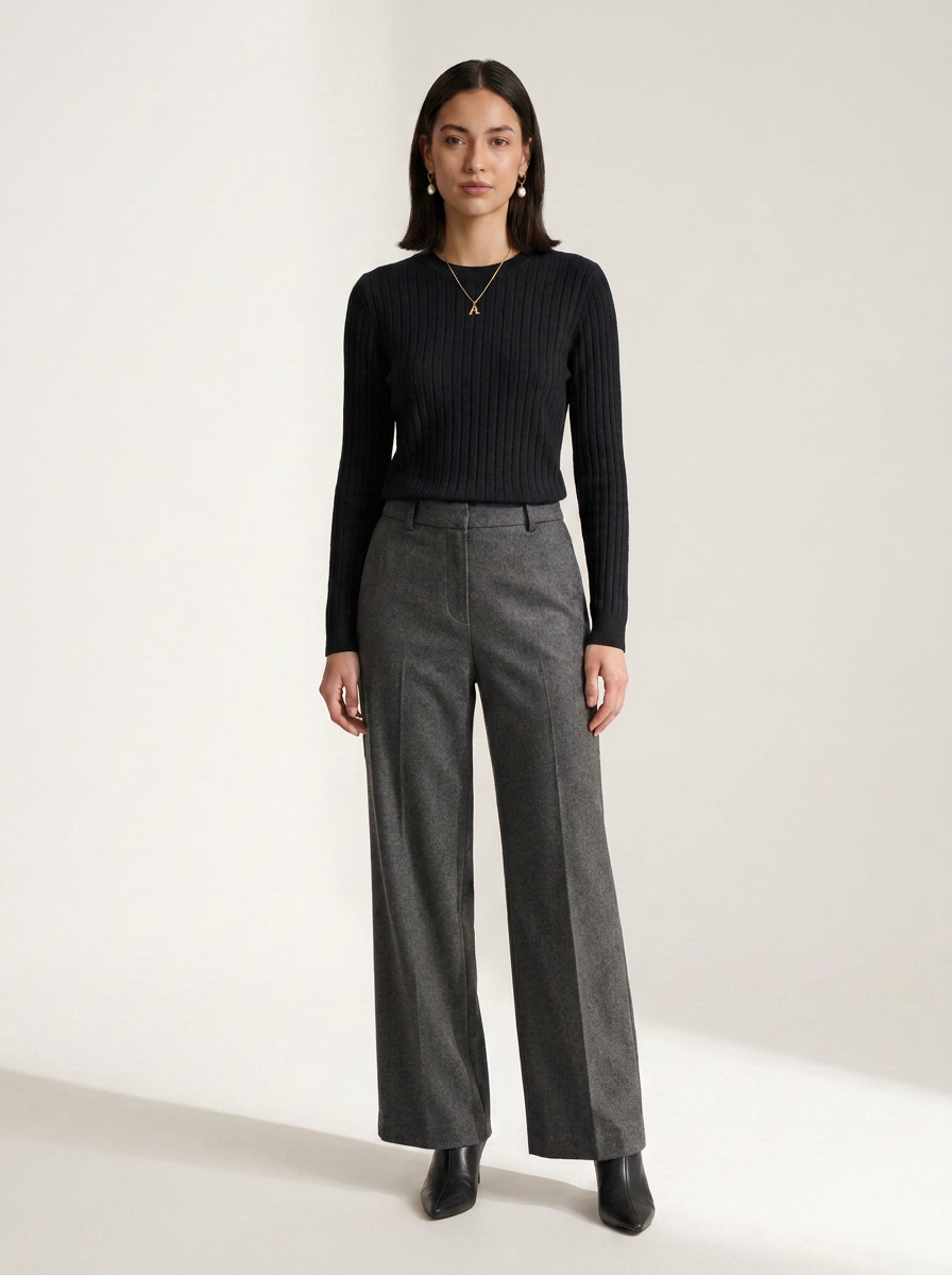 A woman stands against a plain light studio background wearing a black ribbed long-sleeve top and high-waisted gray wide-leg trousers with black ankle boots.