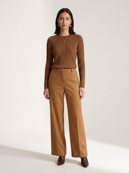 A woman stands against a plain light studio background wearing a fitted ribbed brown long-sleeve top and matching wide-leg brown trousers, with dark pointed shoes.