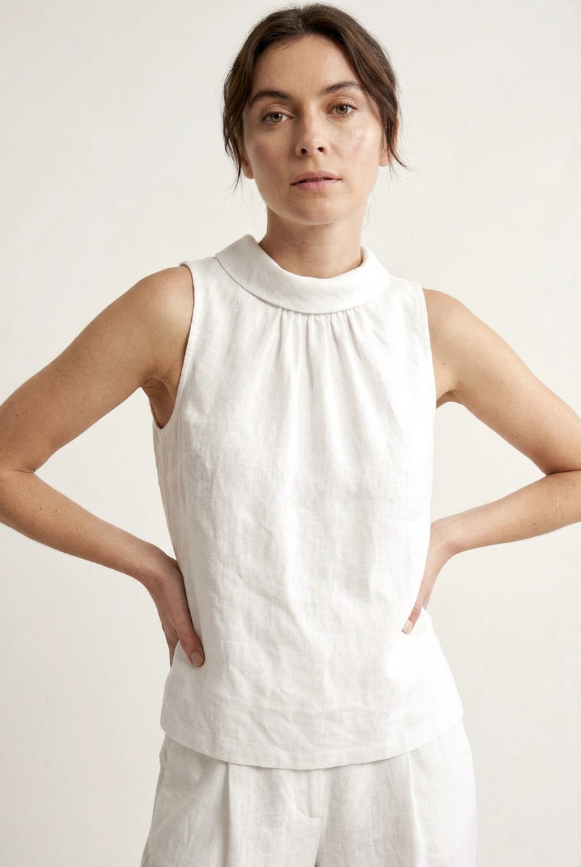 A woman wearing a sleeveless white linen top with a gathered high neck, photographed against a plain light background.