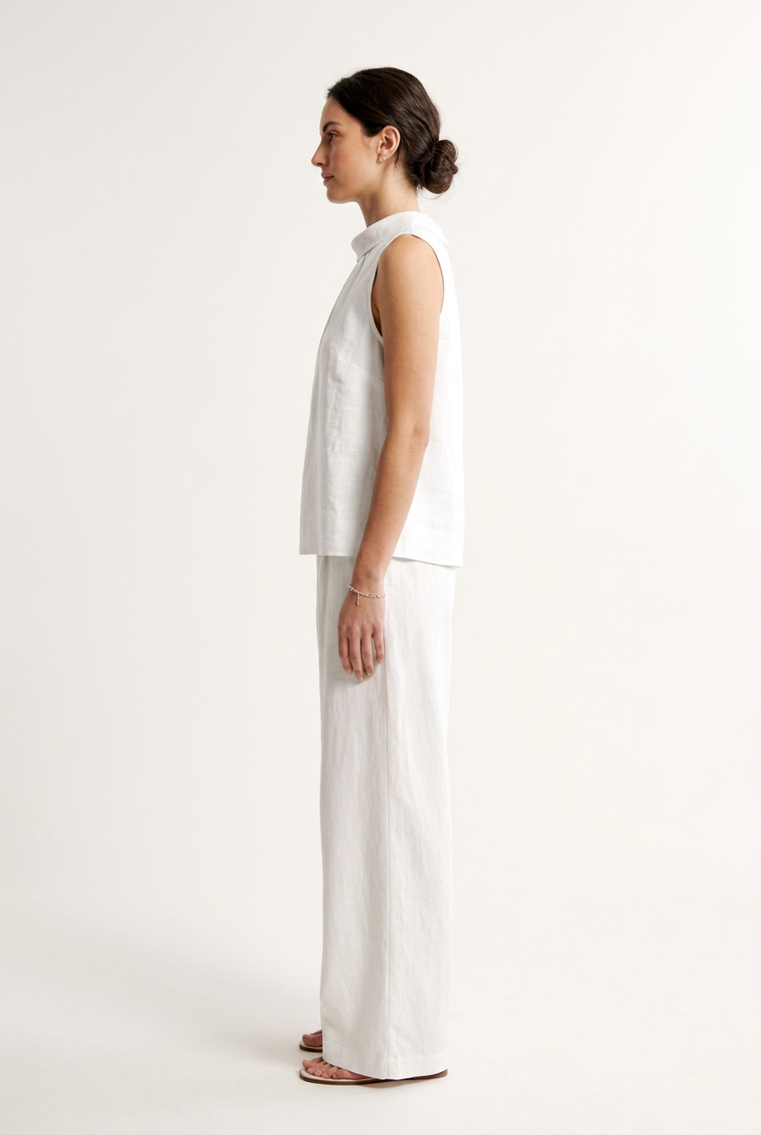 A woman modeling a sleeveless white top with a gathered high mock neck, paired with matching white wide-leg pants, against a plain off-white studio background.
