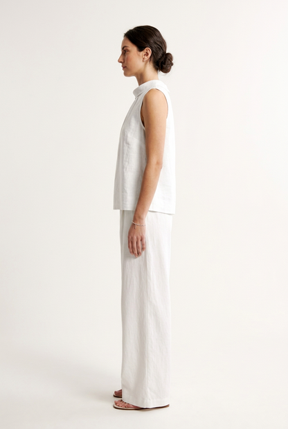 A woman modeling a sleeveless white top with a gathered high mock neck, paired with matching white wide-leg pants, against a plain off-white studio background.