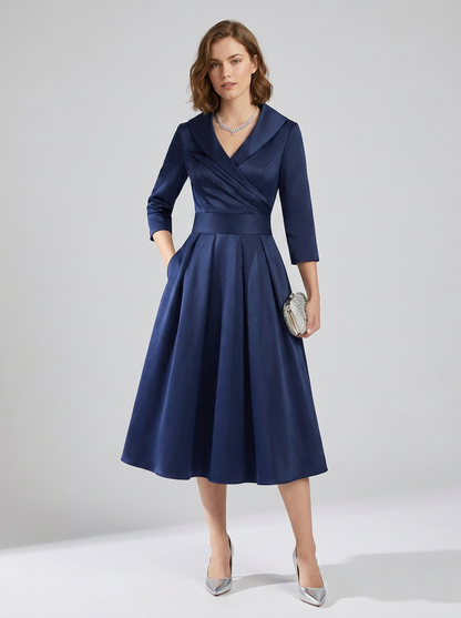 A woman models a navy blue midi dress with three-quarter sleeves, a wrap-style V-neckline, and a flared skirt. She is holding a small silver clutch and wearing silver heels against a light gray studio background.