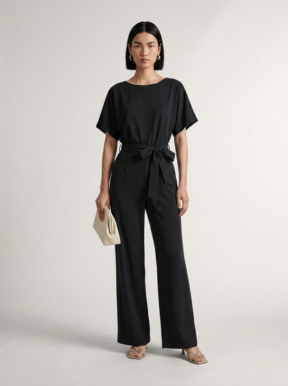 A model stands against a light gray studio background wearing a black short-sleeve belted jumpsuit with wide legs, holding a small cream clutch.