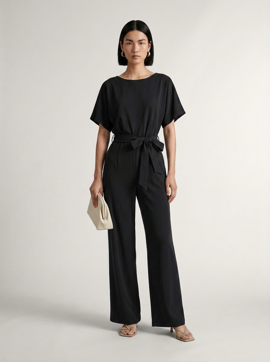 A model stands against a light gray studio background wearing a black short-sleeve belted jumpsuit with wide legs, holding a small cream clutch.