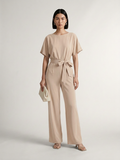 A model stands against a light gray studio background wearing a beige short-sleeve belted jumpsuit with wide legs, holding a small cream clutch.