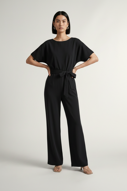 A woman modeling a black short-sleeve jumpsuit with a tie waist and wide legs, standing against a light gray studio background.