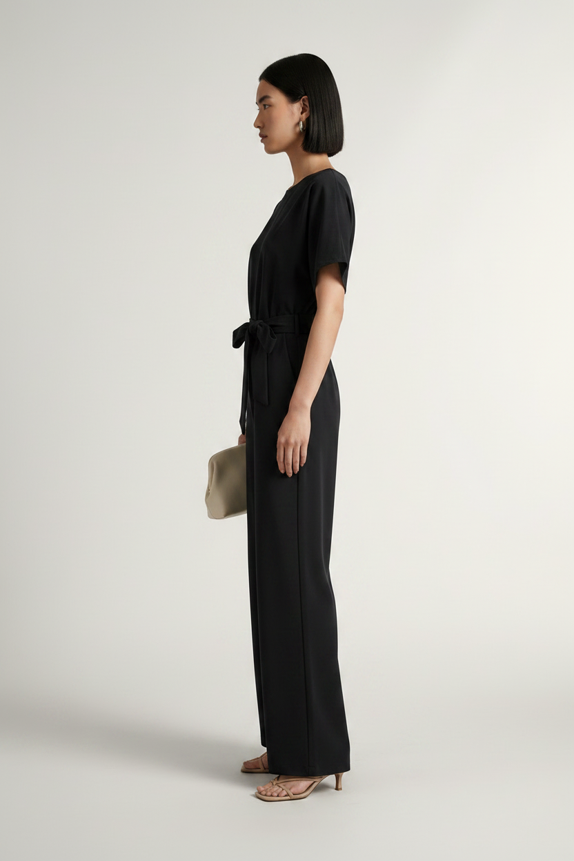 A woman stands in profile wearing a black short-sleeve jumpsuit with a tie waist, paired with nude heeled sandals and holding a small beige clutch, photographed against a plain light gray studio background.