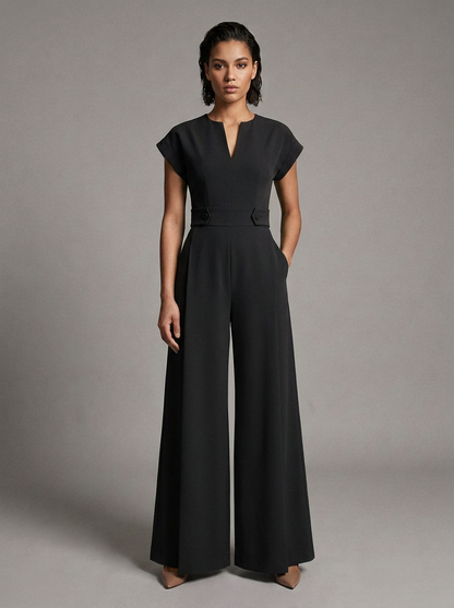 A woman modeling a black wide-leg jumpsuit with short cap sleeves, a split V-neckline, and a matching waist belt, photographed against a neutral gray studio background.