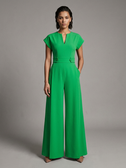 A woman modeling a bright green wide-leg jumpsuit with short cap sleeves, a split V-neckline, and a matching waist belt, photographed against a neutral gray studio background.