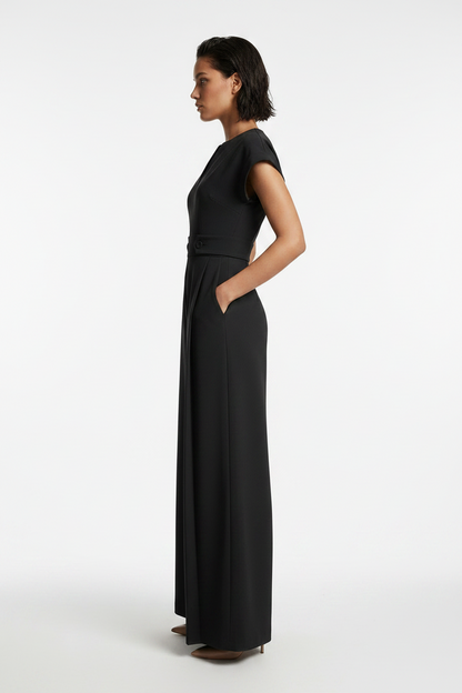A woman modeling a black wide-leg jumpsuit with short cap sleeves, a split V-neckline, and a matching waist belt, photographed against a neutral gray studio background.