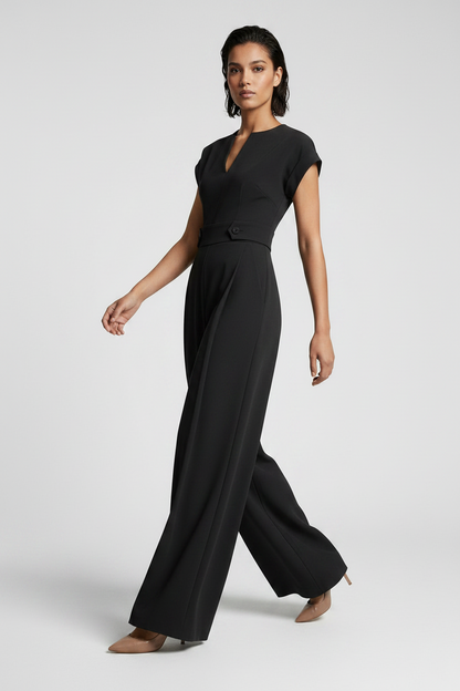 A woman modeling a black wide-leg jumpsuit with short cap sleeves, a split V-neckline, and a matching waist belt, photographed against a neutral gray studio background.