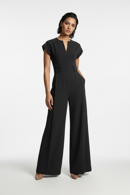 A woman modeling a black wide-leg jumpsuit with short cap sleeves, a split V-neckline, and a matching waist belt, photographed against a neutral gray studio background.