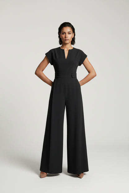 A woman modeling a black wide-leg jumpsuit with short cap sleeves, a split V-neckline, and a matching waist belt, photographed against a neutral gray studio background.