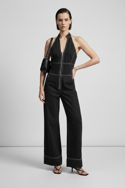 A model stands in a studio wearing a black halter-neck jumpsuit with a front zipper, wide legs, and contrast light piping. She is carrying a small black shoulder bag and wearing black strappy heels against a light gray background.